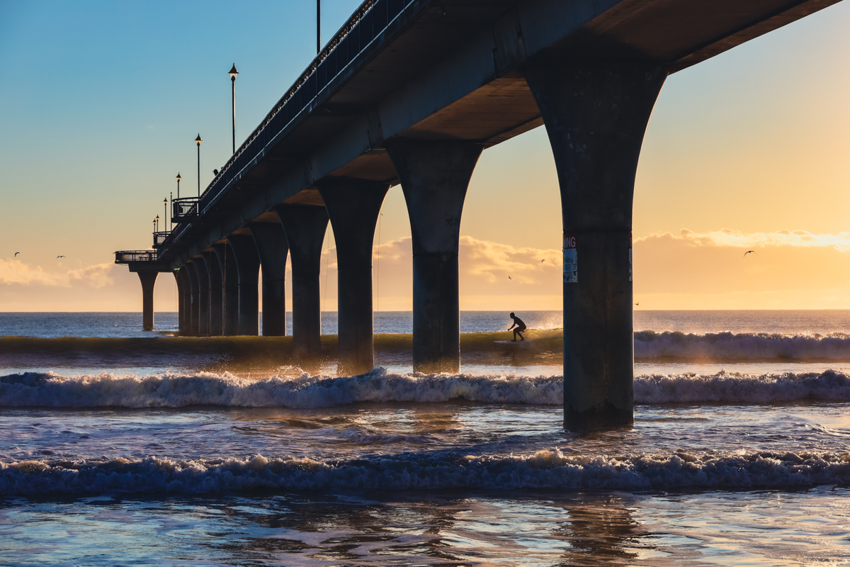 Surfer riding the waves through the New Brighton Pier in Christchurch, New Zealand at sunrise