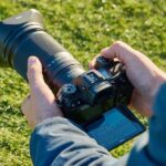 Nikkor X 28-400mm superzoom attached to a Nikon camera photographed over the shoulder of a photographer on a sports field