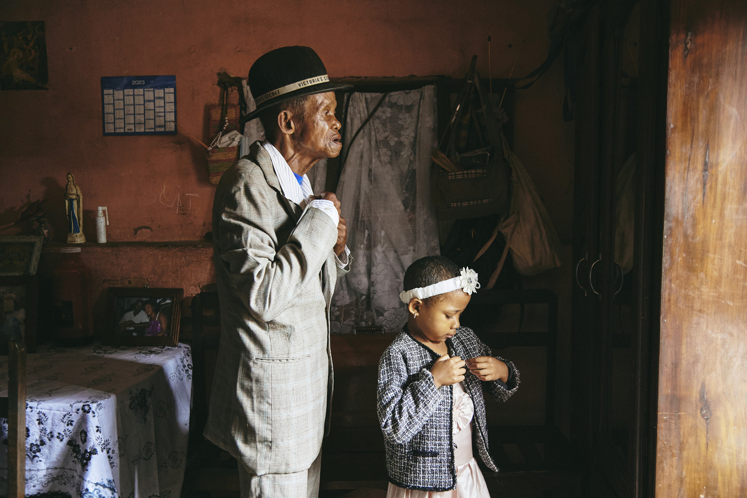 An elderly man with a hat and a young girl with a headband stand in a modestly furnished room. The man is buttoning his jacket while the girl looks down, adjusting her own. A calendar, religious imagery, and family photographs decorate the room, imbuing it with a sense of history and everyday life.