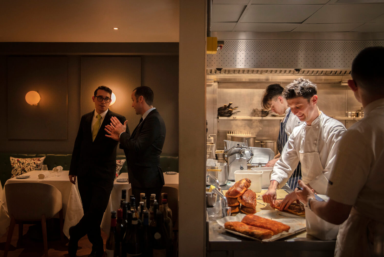 Two men in suits are conversing in an upscale restaurant with a warm and dimly lit ambiance, while chefs are busy preparing food in the open kitchen behind them, adding a sense of behind-the-scenes activity to the dining experience.