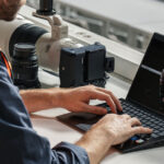 A professional photographer in a high vis press vest is seated at a workstation, reviewing an image on a laptop. Beside the laptop sits a Sony camera. The photographer's focus on the laptop indicates a post-shoot editing or selection process.