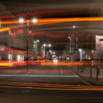 A long-exposure night shot captures the vibrant streaks of light from moving vehicles. The light trails curve through the frame, with city street scenery in the background including illuminated buildings and signage.