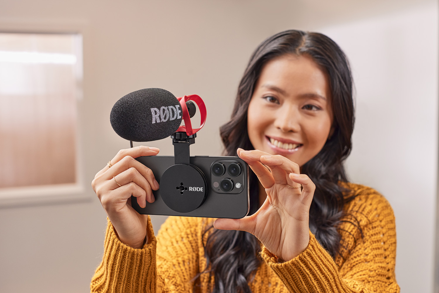 A smiling woman in a yellow sweater is holding a smartphone rig, which includes a RØDE microphone attached to the phone with a black circular magnetic mount. The setup is designed for mobile content creation, with the microphone prominently featuring the Rode logo.