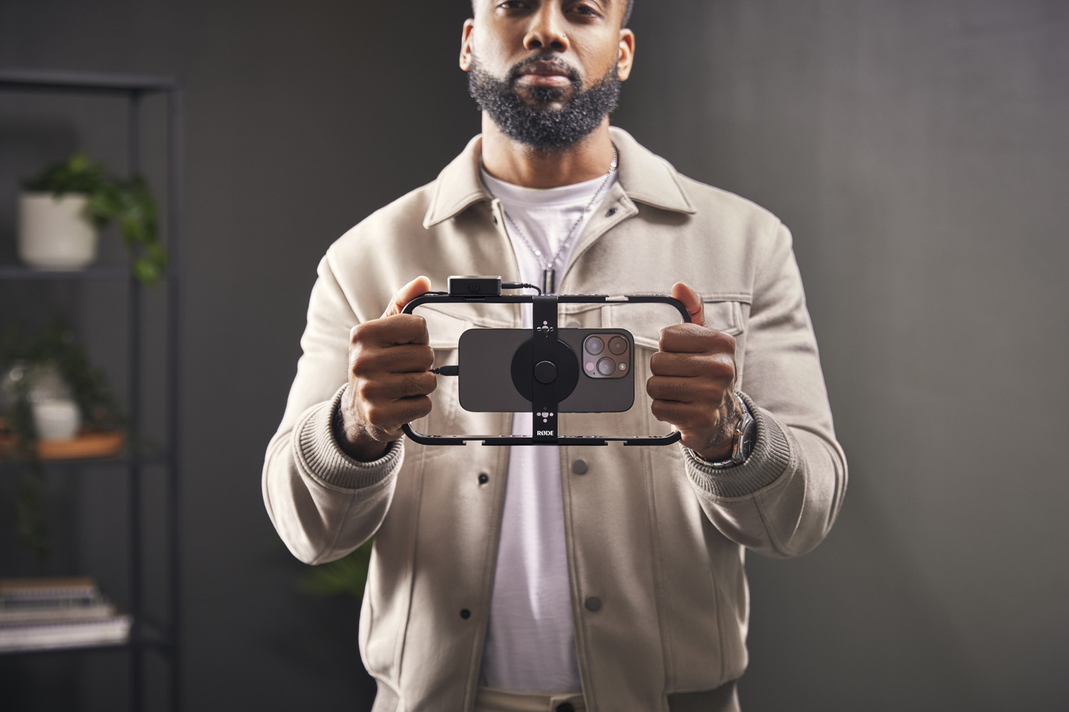 A focused man in a light beige jacket holds a RØDE Phone Cage with a smartphone mounted in the center. The cage has a central circular RØDE mount with the company's logo, designed to attach various filmmaking accessories. The man's confident stance suggests readiness to begin content creation.