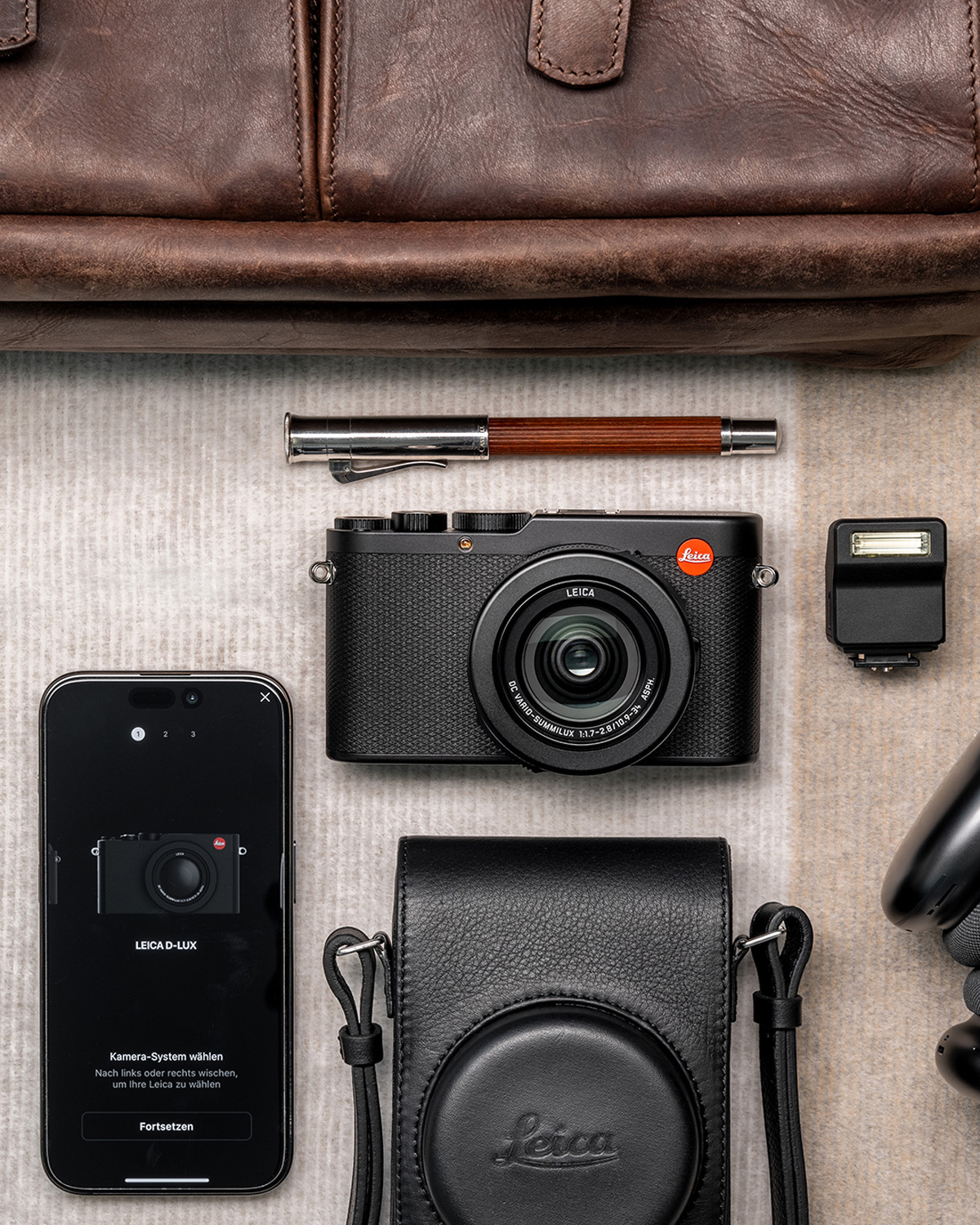 Product lifestyle flat lay image of Leica D-Lux 8 arranged alongside a leather back, leather case, pen, show mount flash and smartphone