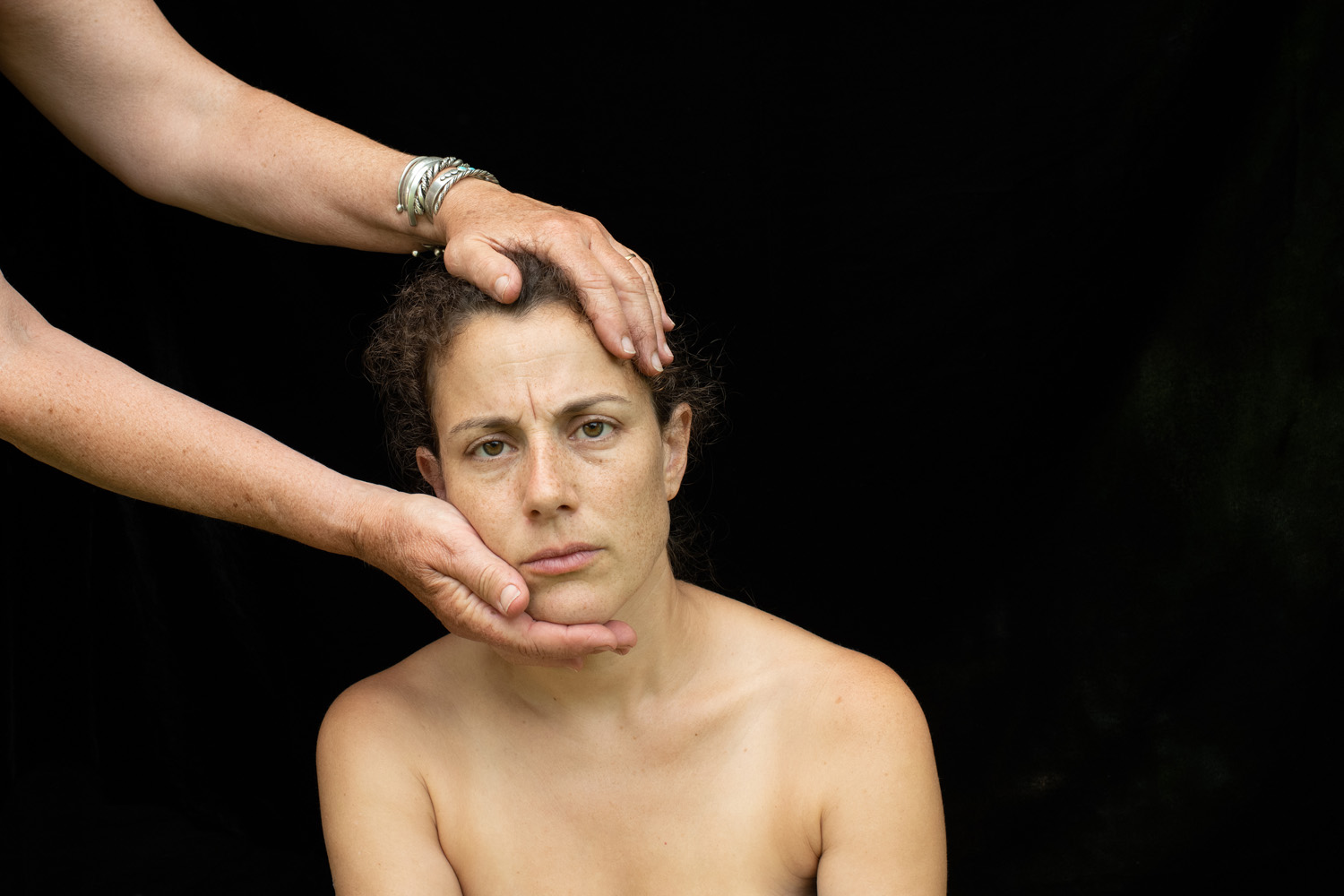 A close-up portrait of a woman with a solemn expression, bare shoulders, looking directly into the camera. Two hands, one on her forehead and one supporting her chin, frame her face against a dark background.