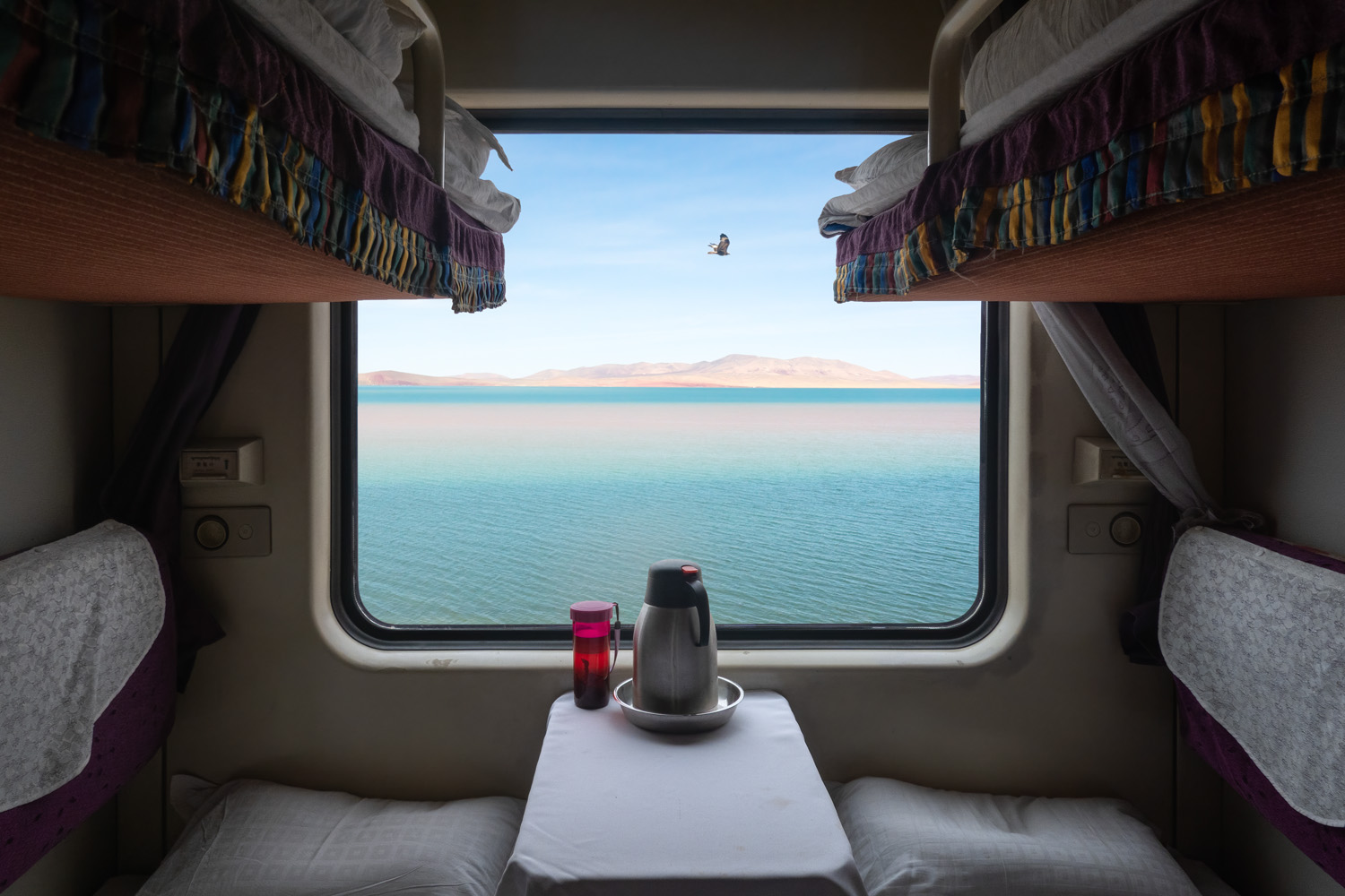 A peaceful view from a train cabin window, showing a vast lake with clear blue water and distant mountains. The interior includes two bunks, a table with a thermos, and a red cup.