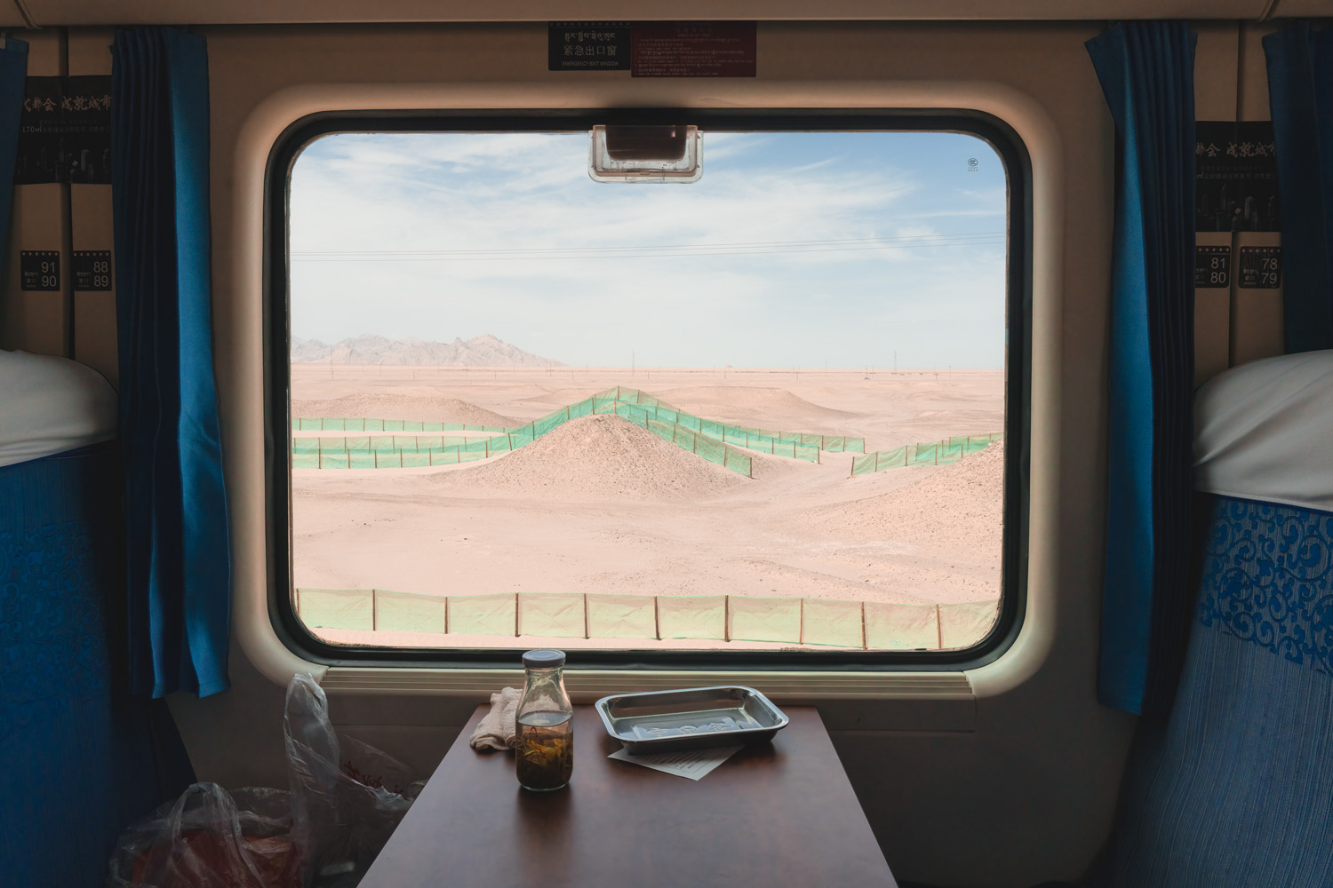 A desert landscape viewed from a train window, featuring rolling sand dunes and green fencing. The scene is framed by the interior of the train cabin.