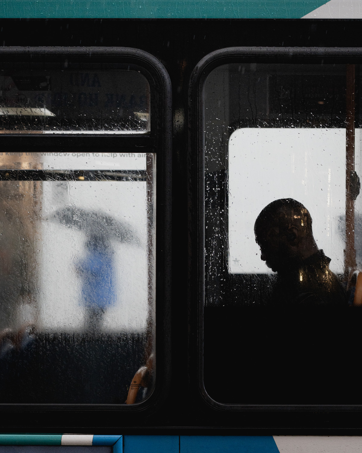 A view through a rain-speckled bus window showing a silhouette of a man sitting inside, while outside a person with an umbrella is walking past.