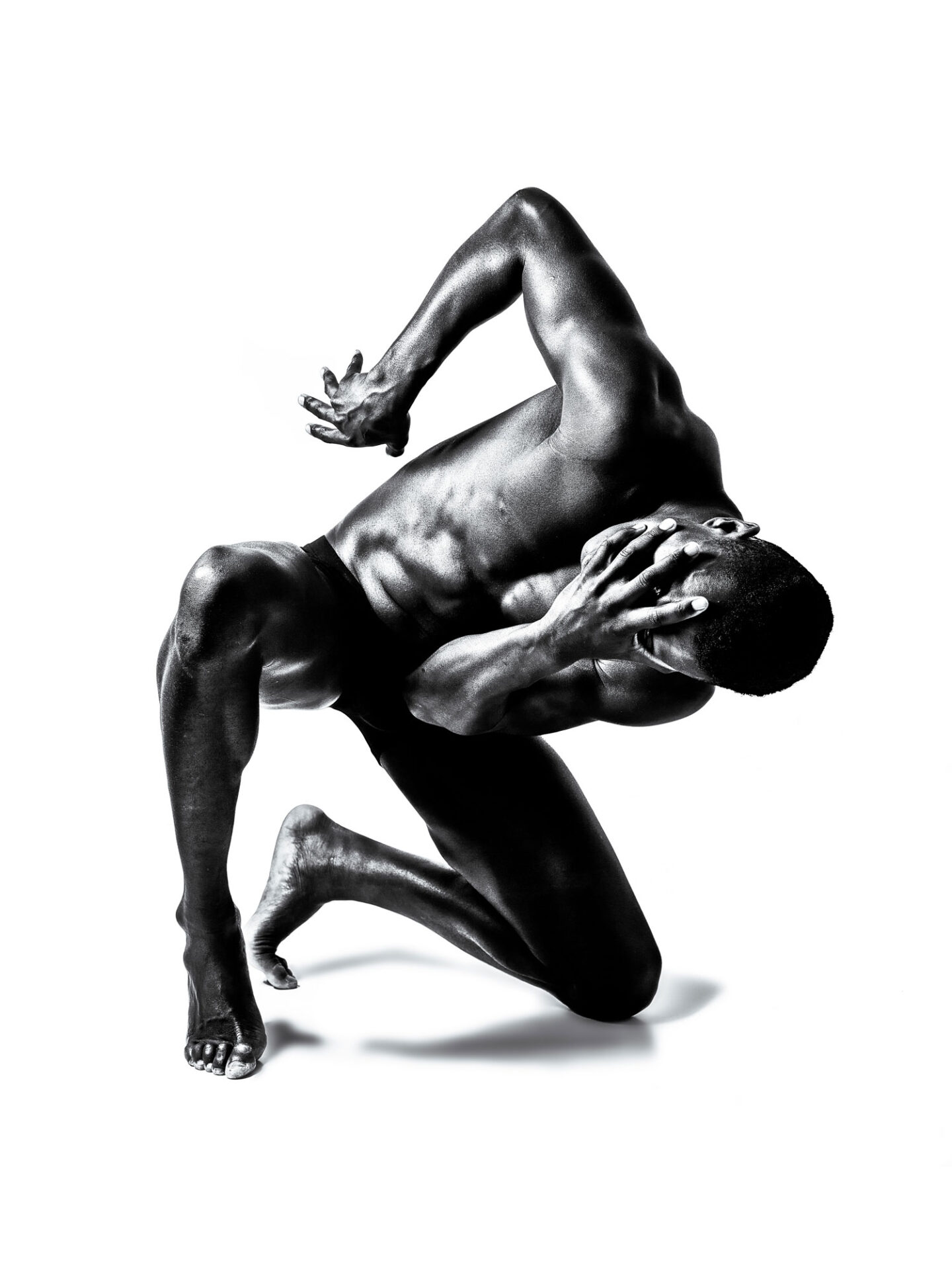 A monochrome photograph of a muscular man in a dynamic, twisted pose, with one hand over his face and the other arm outstretched, set against a plain white background, showcasing the tension and movement in his body.