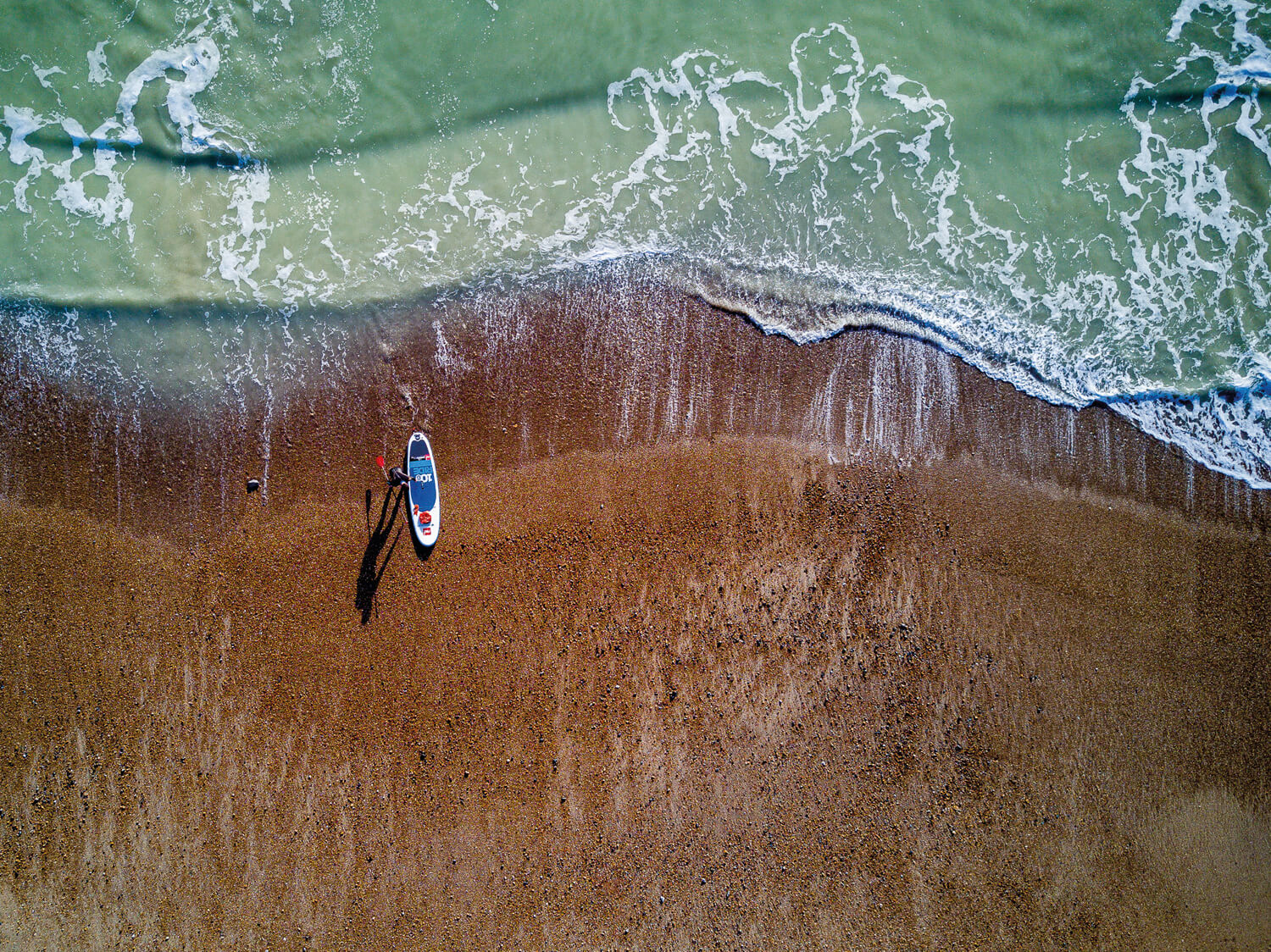 A top-down photograph of a beach where a lone paddleboarder stands next to a paddleboard. The board is positioned near the edge of the water, and the waves gently lap at the shore, creating a contrast between the blue water and the brown sand.