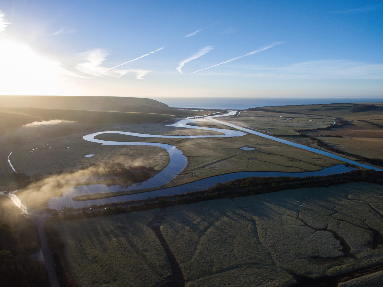 An aerial photograph showcasing a winding river meandering through a flat landscape. The sun casts long shadows across the fields and water, and the scene is framed by a distant horizon and a clear sky.