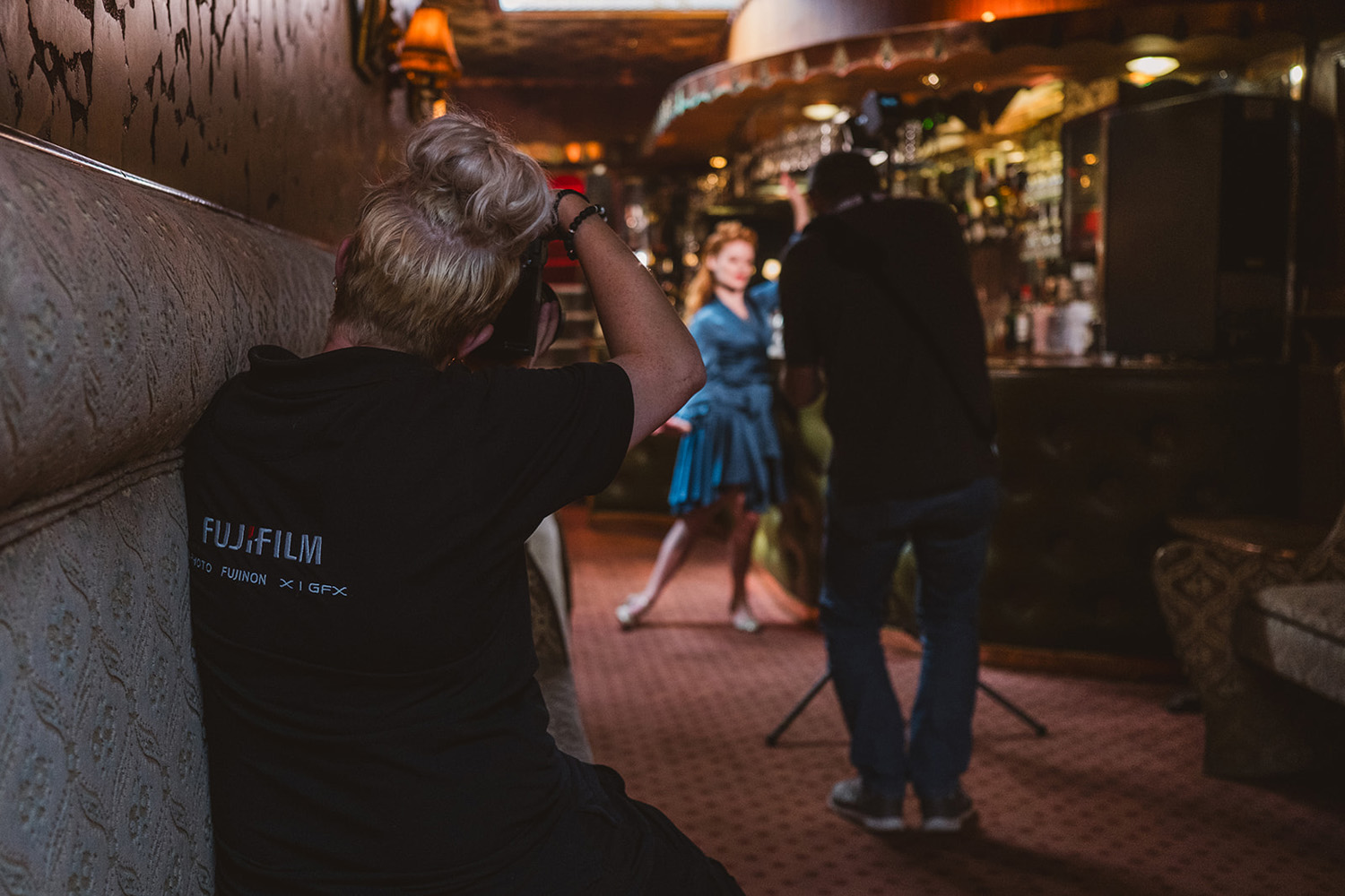 A photographer with blonde hair is using a Fujifilm camera to photograph a model posing in a dimly lit, ornate bar. Another photographer is adjusting lighting equipment in the background.