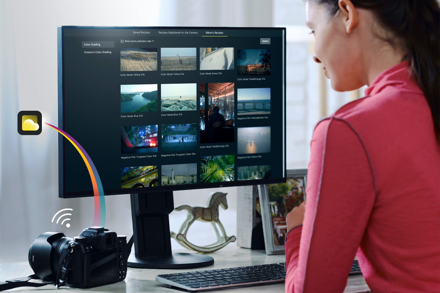 A woman is sitting at a desk using a desktop computer. The monitor displays a screen with various photo thumbnails. A camera is connected to the computer, and an icon showing a cloud with a Wi-Fi signal illustrates the data transfer from the camera to the computer. The woman appears to be editing or organising photos.