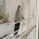 A woman stands on the balcony of an apartment building, framed by an intricate metal railing design. She looks at the camera pensively, with the facade of another building and some greenery visible in the background.