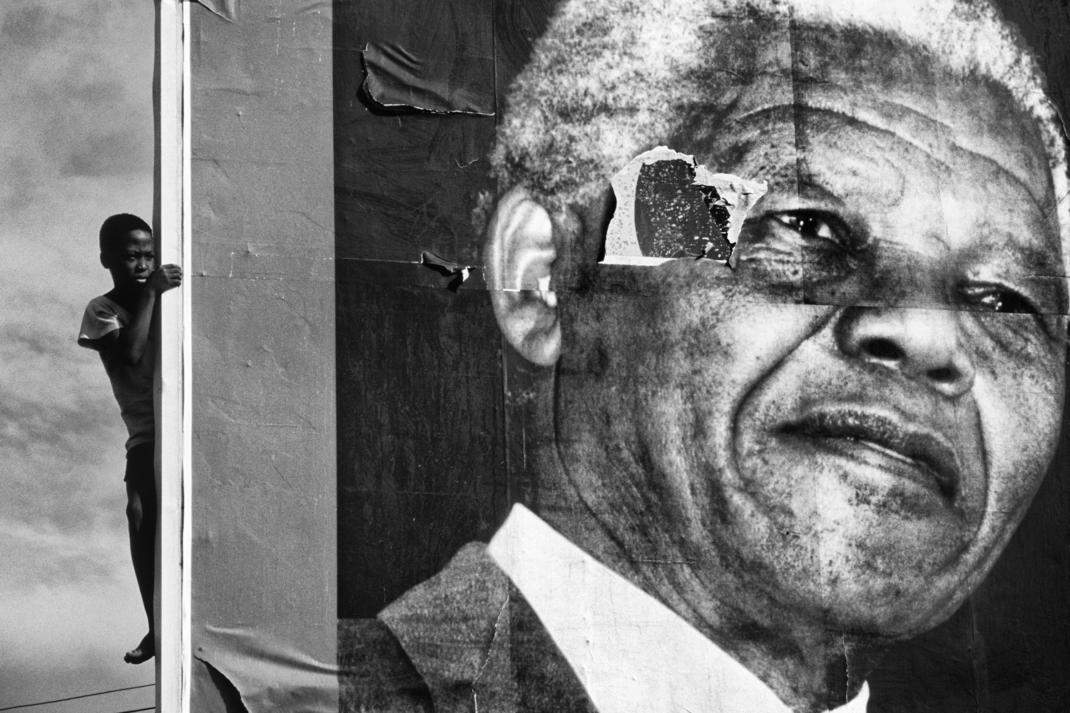 Black and white photograph of a young boy clinging to the side of a large, weathered poster of Nelson Mandela. The torn and worn sections of the poster give it a textured, powerful feel.