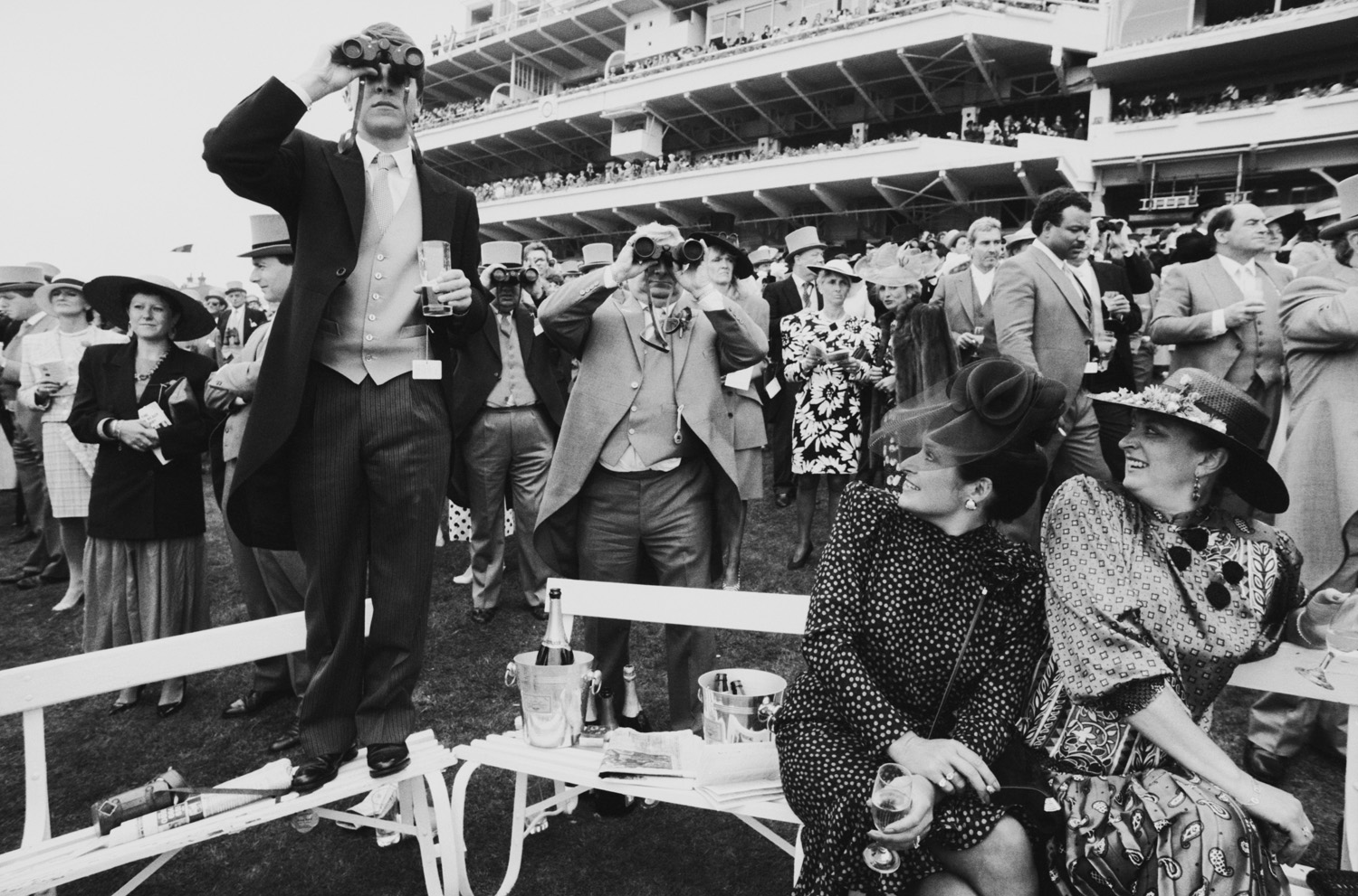 Black and white photograph of spectators at a horse racing event, dressed in formal attire, with one man standing on a bench and looking through binoculars, capturing the elegance and anticipation of the moment.