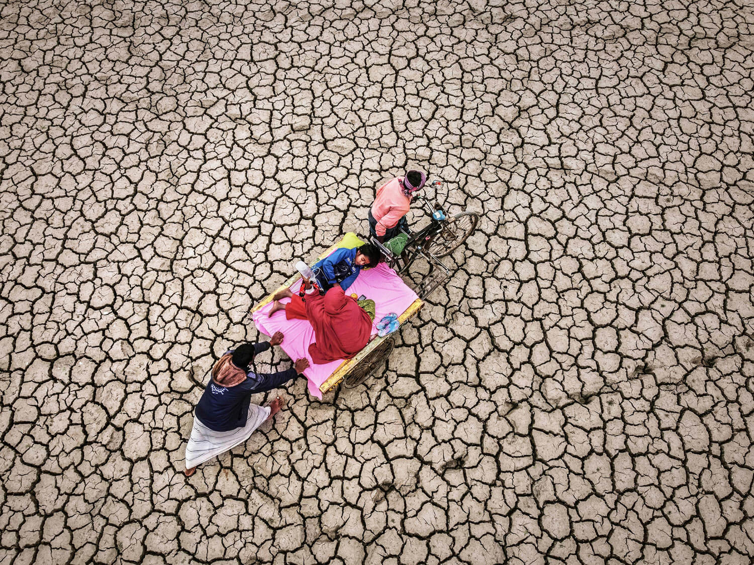 An overhead view of a man pushing a rickshaw carrying a family across a vast, cracked, drought-affected landscape, symbolising the impact of climate change on communities reliant on the land.