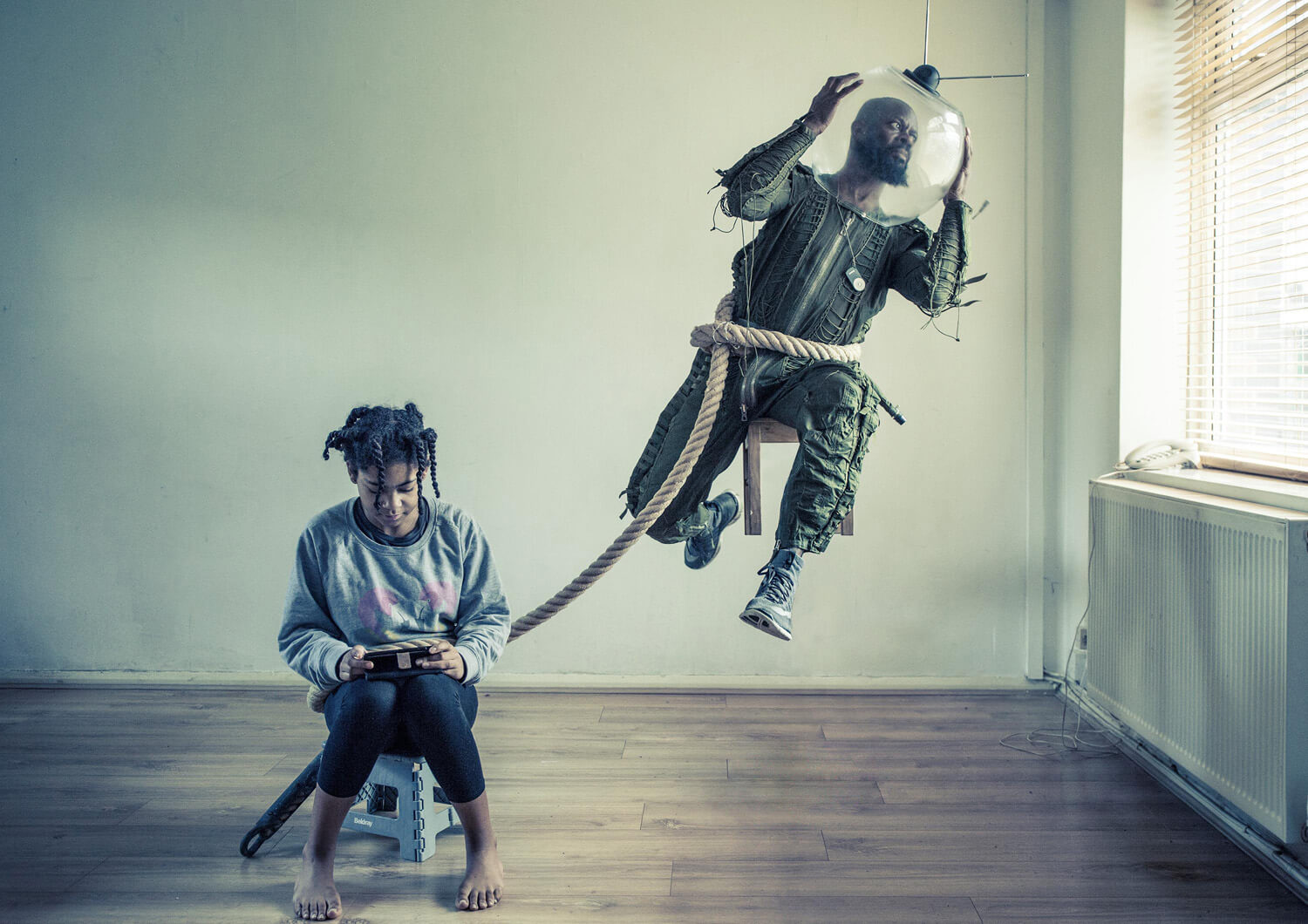 A surreal indoor scene with a young girl sitting on a stool, looking at a tablet, while a man in a spacesuit-like helmet is tethered by a rope, highlighting the themes of mental health and isolation.